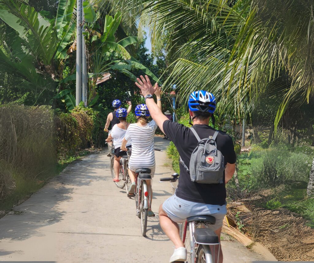 Four travelers cycling through a peaceful countryside road in Ben Tre, Vietnam, waving hands joyfully.