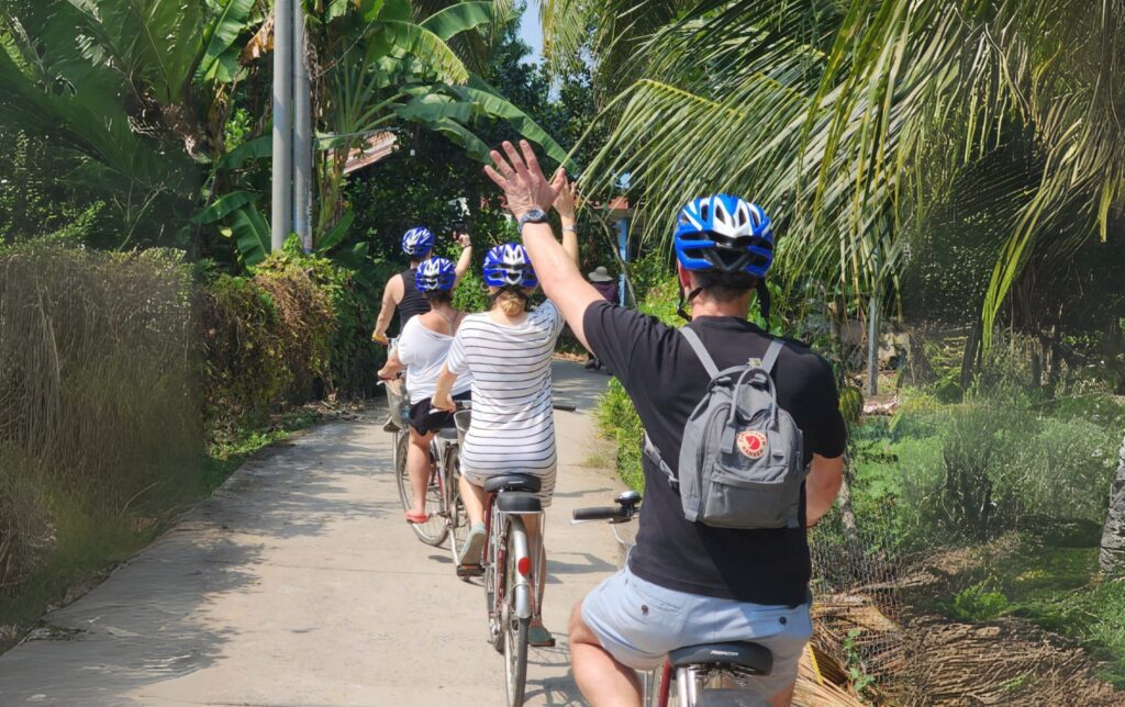 Four travelers cycling through a peaceful countryside road in Ben Tre, Vietnam, waving hands joyfully.