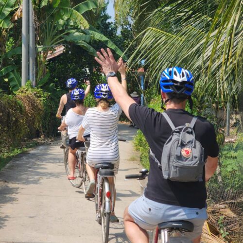 Four travelers cycling through a peaceful countryside road in Ben Tre, Vietnam, waving hands joyfully.
