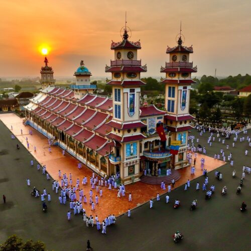 Sunset view of Cao Dai Temple in Tay Ninh, with worshippers dressed in traditional white robes gathered around the colorful temple