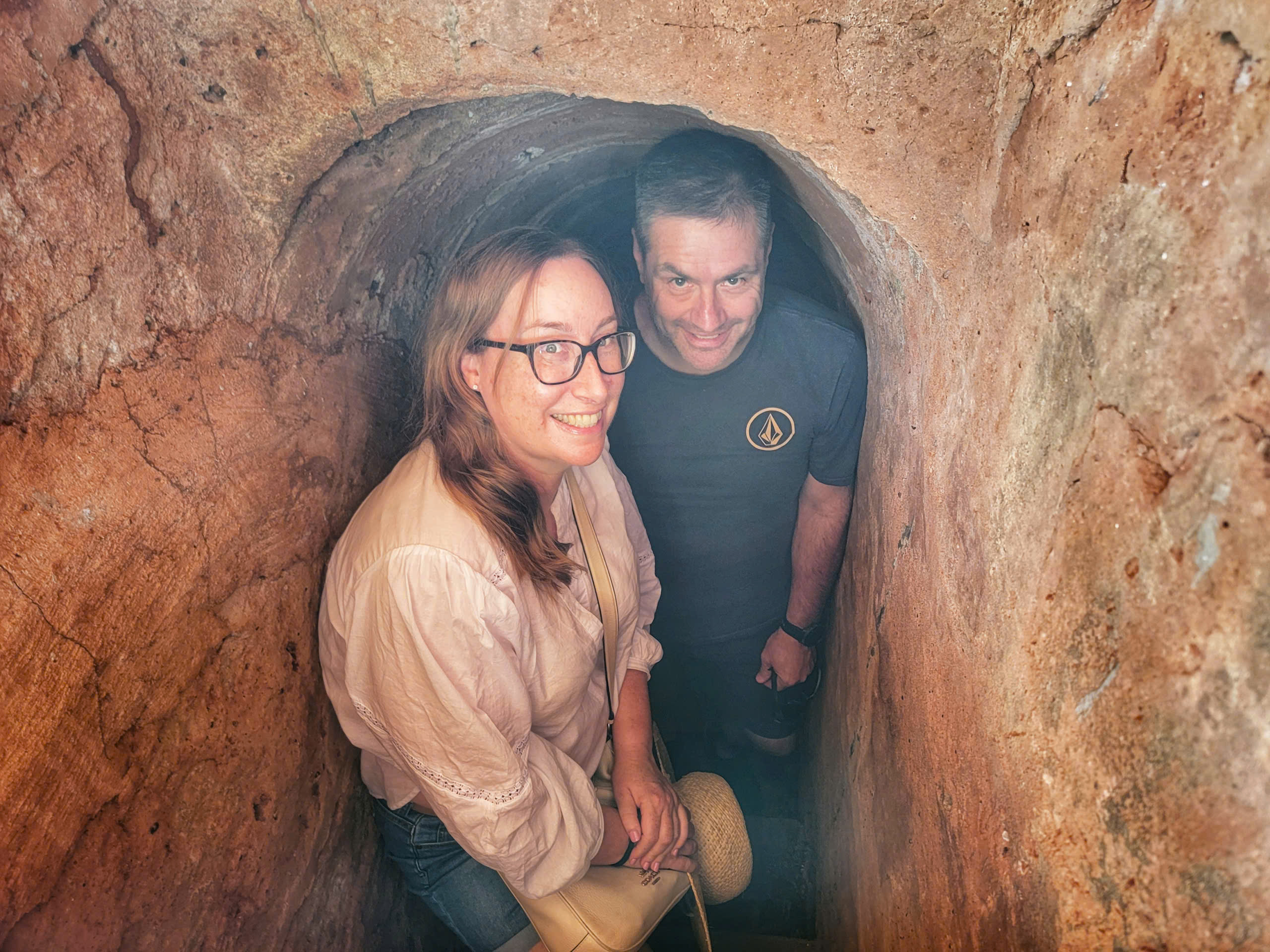 Two visitors smiling inside the narrow underground tunnel at Cu Chi.