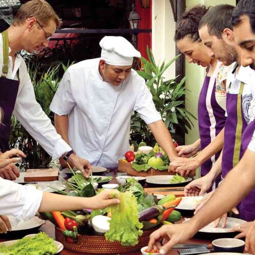 A chef guiding participants around a table during a Saigon cooking class, with fresh green vegetables prepared for traditional Vietnamese dishes.