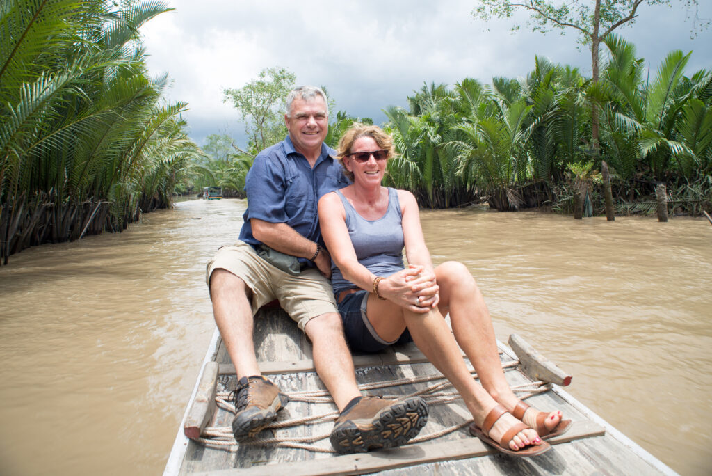 Two travelers taking photos on boat amid nipa palm river scenery.