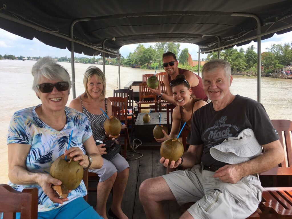 5 tourists are sitting on a boat on the river in mekong delta and happily drinking whole coconut water