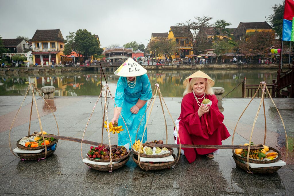 Two female travelers wearing traditional Vietnamese ao dai and conical hats posing with a fruit vendor, with the historic streets of Hoi An Old Town in the background, capturing the soul of central vietnam heritage.