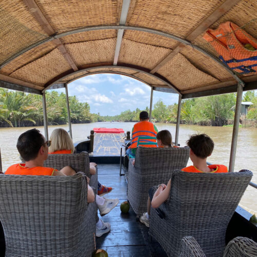 **Alt text chuẩn SEO đề xuất:** Travelers wearing life jackets seated on a motorized boat cruising through nipa palm-lined waterways in the Mekong Delta