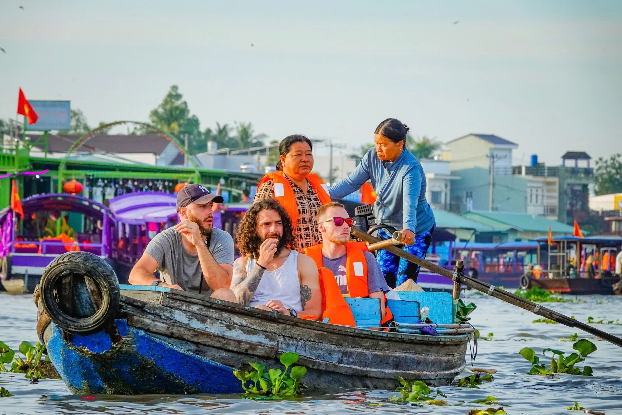 Three tourists on boat tour through floating market rowed by local women.
