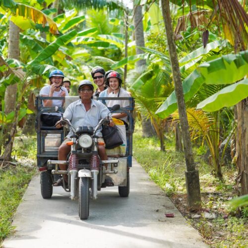 Local driver riding a xe lôi (tuk-tuk) with smiling travelers along a narrow village road lined with coconut and banana trees in Ben Tre, Mekong Delta.