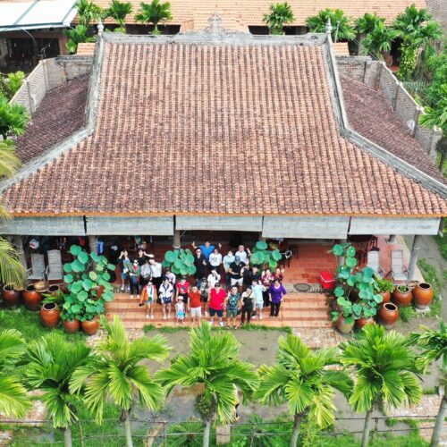 Top-down view of a Mekong Delta homestay with tiled roof and locals warmly greeting visitors.