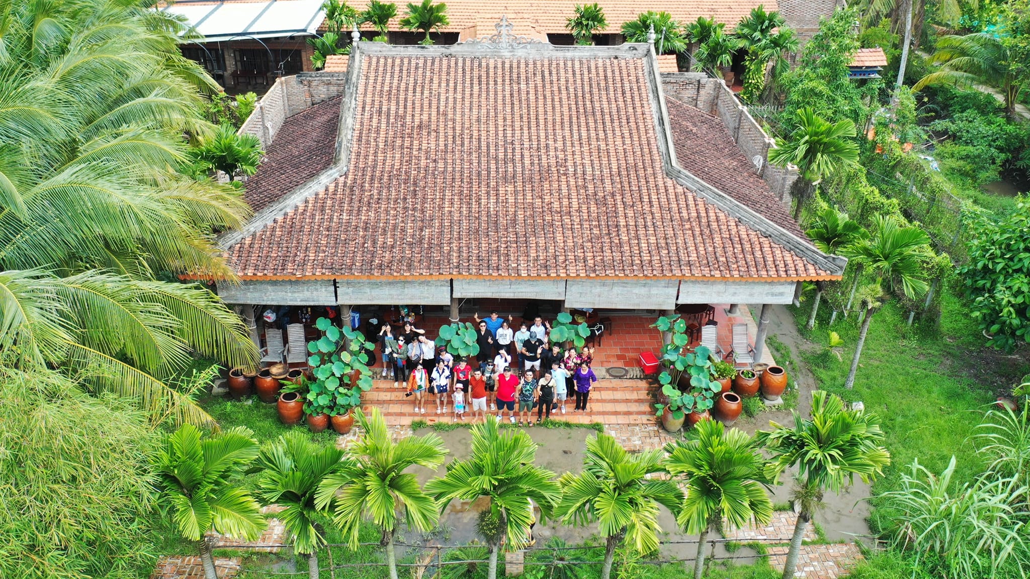 Top-down view of a Mekong Delta homestay with tiled roof and locals warmly greeting visitors.