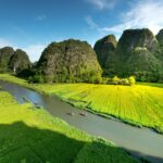 Aerial view of Tam Coc Ninh Binh river with two small boats gliding between lush green banks, framed by dramatic limestone karst mountains in the background.