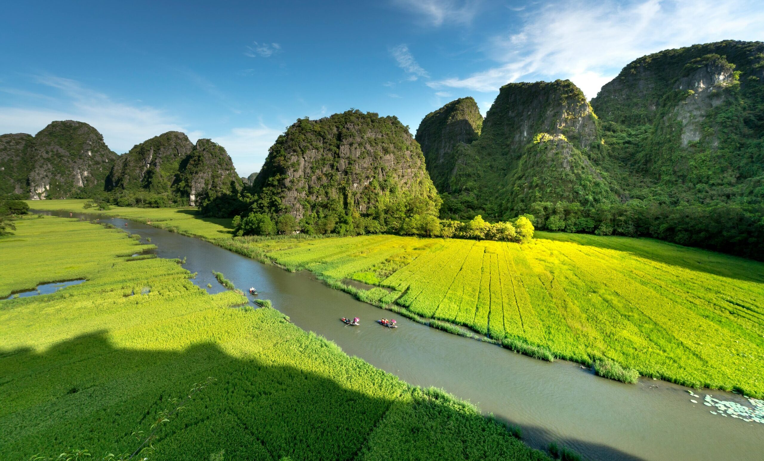 Aerial view of Tam Coc Ninh Binh river with two small boats gliding between lush green banks, framed by dramatic limestone karst mountains in the background.