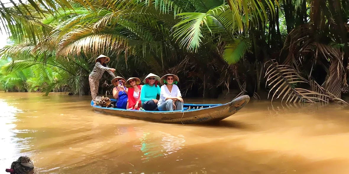 A local woman rows a sampan carrying smiling travelers in conical hats along a quiet river lined with nipa palms in Ben Tre, offering an authentic Mekong Delta experience.