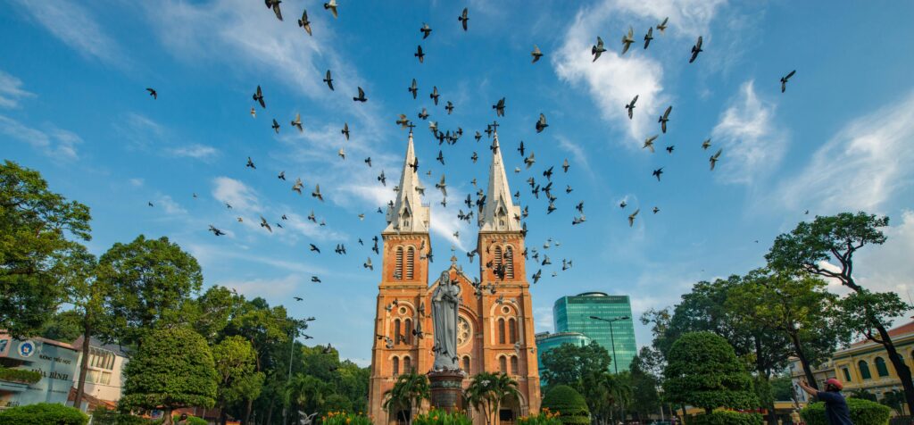 Symmetrical front view of Saigon Notre Dame Cathedral with a flock of pigeons flying across the blue sky