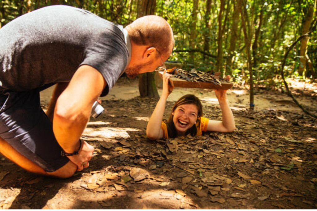 A man watching a smiling young girl crawl into the narrow Cu Chi Tunnels, experiencing wartime history on a Cu Chi Tunnels tour in Vietnam.