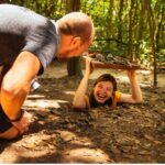 A man watching a smiling young girl crawl into the narrow Cu Chi Tunnels, experiencing wartime history on a Cu Chi Tunnels tour in Vietnam.