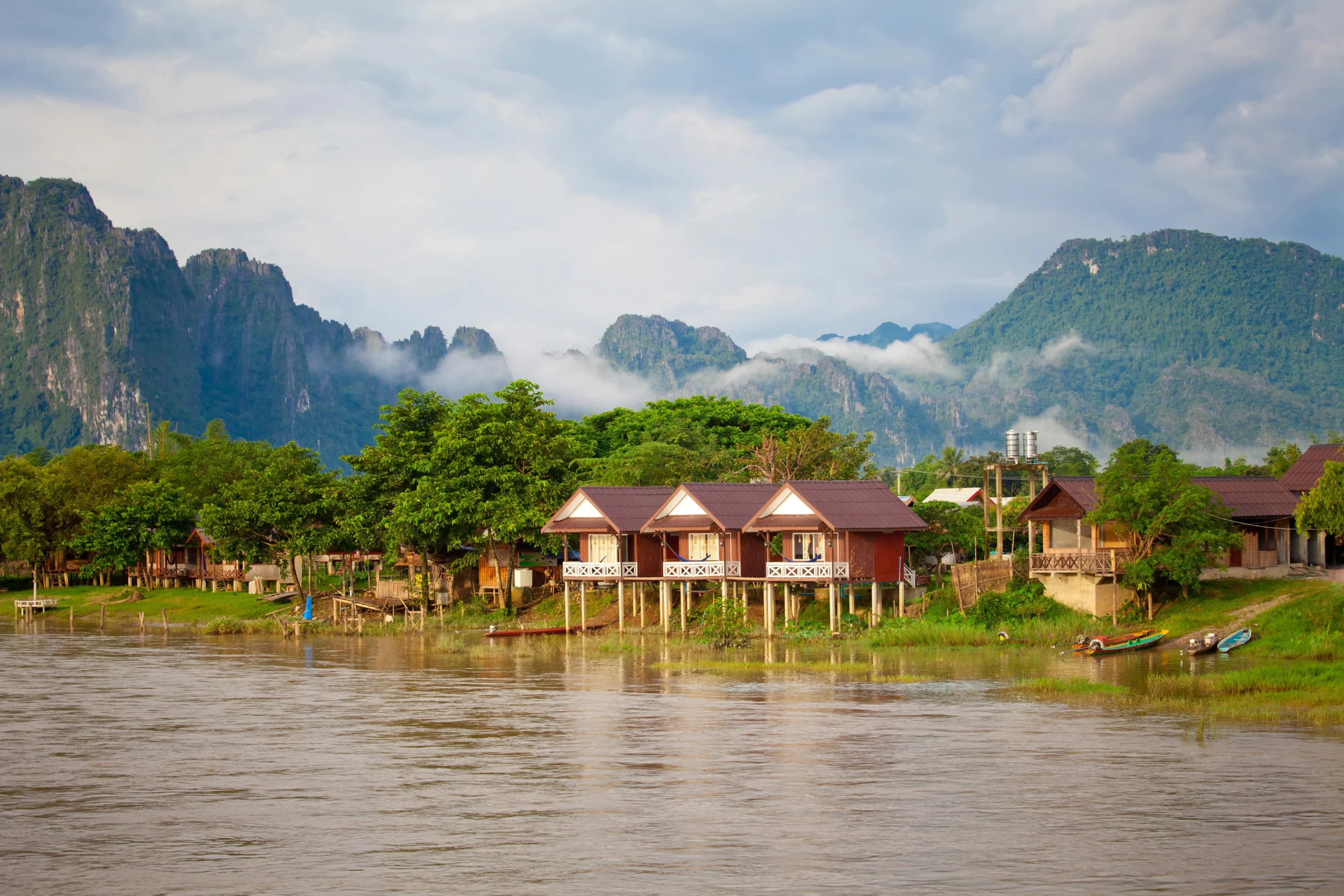 Traditional red-roof stilt houses lining the river in Vang Vieng town, Laos, with dramatic limestone mountains rising in the background.